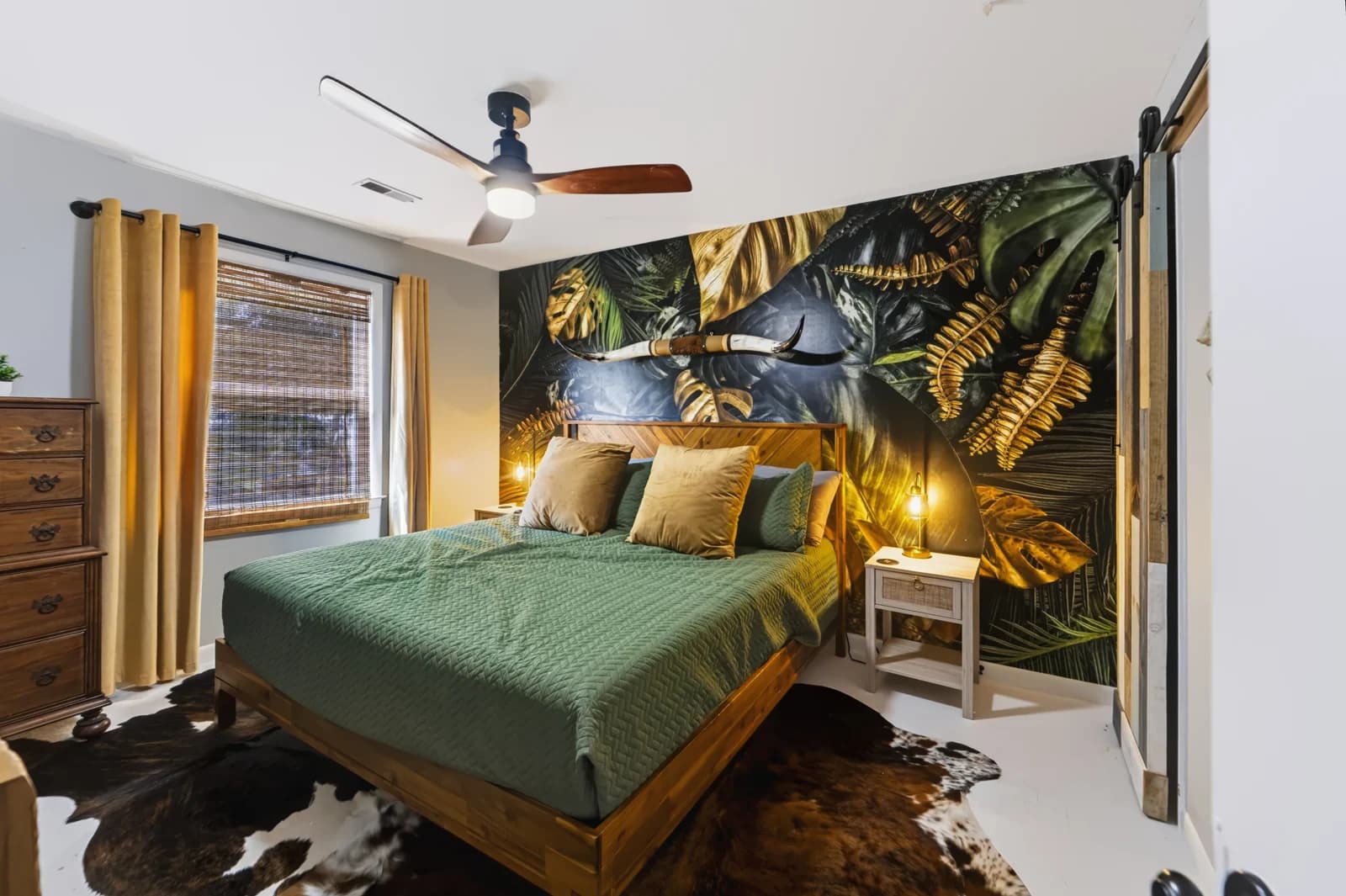 Bedroom with green bedding, botanical wallpaper, wood furniture and warm bedside lighting