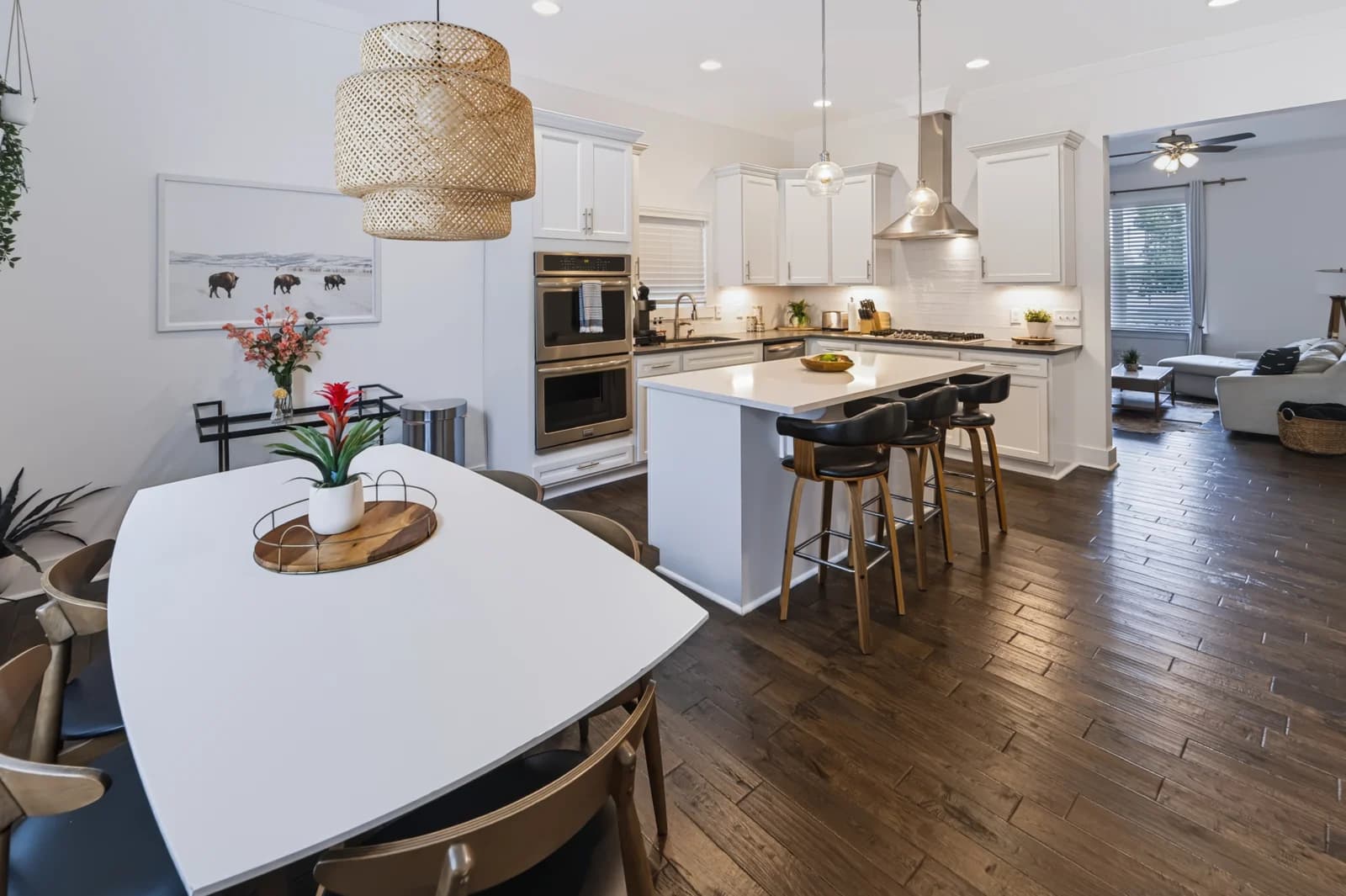 Dining table beneath a woven pendant looking toward a white kitchen and living room
